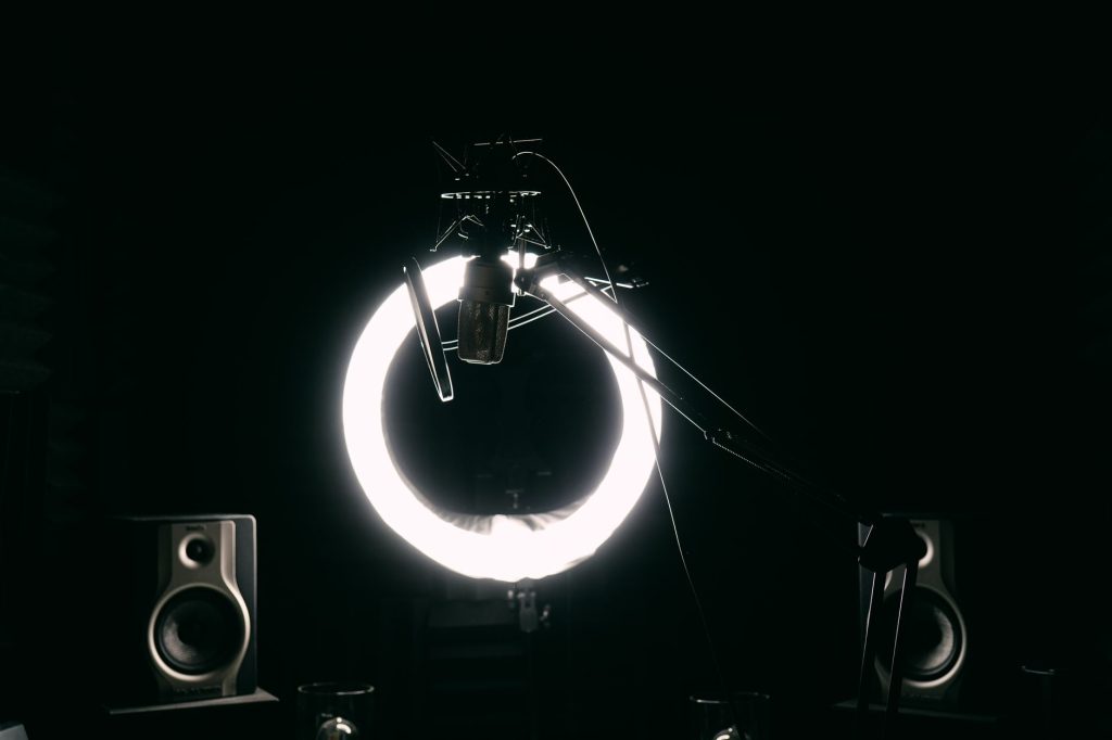 Dark studio room with condenser microphone, ring light, and speakers for recording purposes.