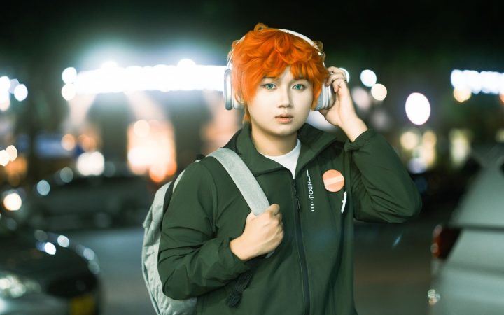 Young adult with striking orange hair and headphones in a nighttime urban setting.