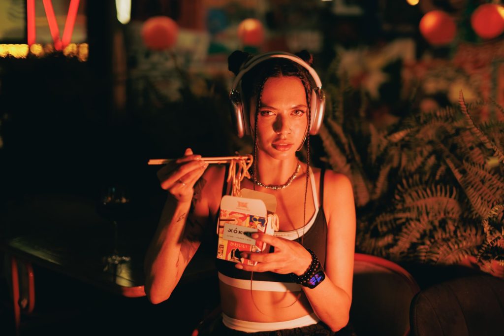 Stylish woman with headphones eating noodles outdoors under neon lights at night.