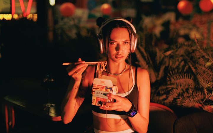 Stylish woman with headphones eating noodles outdoors under neon lights at night.